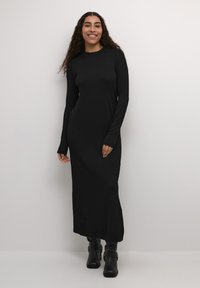 Black long-sleeve maxi dress in soft fabric with a fitted silhouette. Features a round neckline and thumbholes at the cuffs. Worn with black ankle boots.