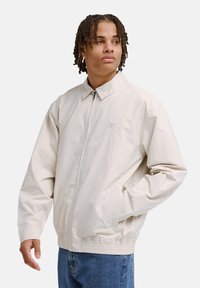 Giacca leggera beige con colletto classico, cerniera frontale, orlo elastico e tasche laterali, con un logo discreto sul petto.