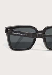 Ochelari de soare din acetat negru cu ramă pătrată, lentile gri și logo-ul "Armani Exchange" pe templu. Textură netedă și design îndrăzneț.