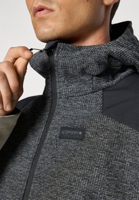 Gray textured zip-up jacket with a hood, featuring a black shoulder panel and a logo label on the front, detailing a snug fit.