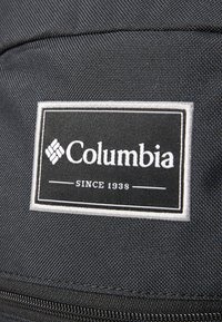 Fekete szövet hátizsák, téglalap alakú Columbia logóval, fehér szöveggel és kontúrral, amelyen a "SINCE 1938." felirat szerepel. Texturált felület.