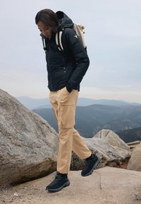 Black insulated jacket, beige cargo pants, and black hiking shoes on rocky terrain. A beige backpack is worn, with a natural landscape in the background.