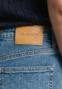 Donkerblauwe geribde top boven lichte blauwe denim jeans, met een tan leren label met het reliëf "GANT" logo en zichtbare stiksels.