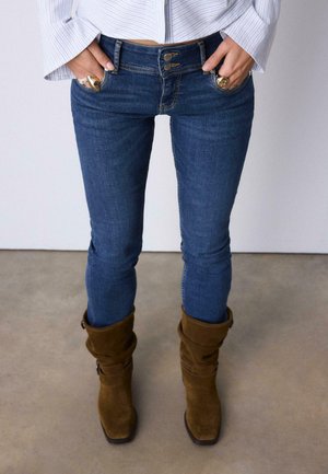 Persoon draagt blauwe skinny jeans met dubbele knoopsluiting, wit gestreept shirt, bruine suède laarzen en gouden ringen aan beide handen.