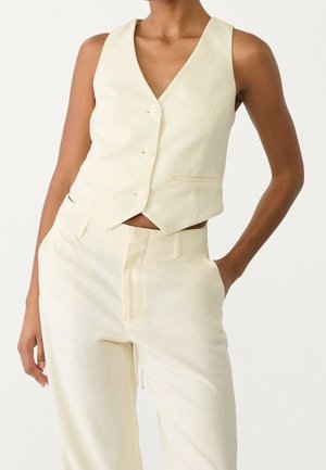 Vrouw met mouwloze crèmewitte gebuttonde gilet en bijpassende hoge taille broek, met één hand in de zak, tegen een effen achtergrond.