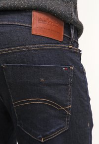 Jeans de mezclilla azul oscuro con una etiqueta de cuero marrón que presenta "HILFIGER DENIM" grabado, costuras amarillas en contraste y un diseño en el bolsillo trasero.