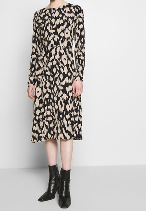 Mujer con un vestido beige y negro de manga larga y largo hasta la rodilla con un patrón abstracto, y botas negras hasta el tobillo, sobre un fondo blanco liso.