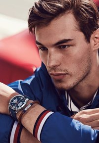 Orologio argento con quadrante blu e marcatori delle ore prominenti, indossato su un polso con un cinturino in pelle marrone. Giacca blu con accenti rossi e bianchi.