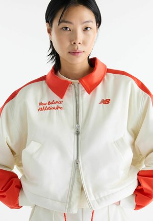 Ung person iført hvid og orange New Balance Athletics lynlåsjakke, stående med hænderne i lommerne mod en hvid baggrund.