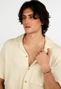 Camicia button-up corta di colore crema, con colletto rilassato e una sottile trama. Il modello indossa un bracciale metallico su un polso.