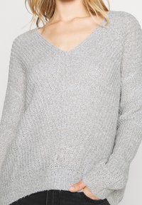 Pull en tricot gris clair avec un col en V, manches longues, motif texturé et ourlet côtelé. Présente une coupe décontractée et un design en tricot ouvert.