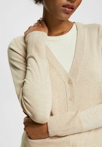 Beżowy kardigan z miękkiej dzianiny o dekolcie w kształcie litery V, z czterema małymi guzikami i ściągaczami przy rękawach, w połączeniu z jasnym topem.