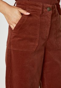 Pantaloni in corduroy color ruggine con tasca laterale, linee verticali testurizzate e chiusura con bottone in vita.