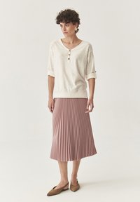 Helles cremfarbenes Strickoberteil mit kurzen umgeschlagenen Ärmeln und Knopfdetails, kombiniert mit einem plissierten, knielangen rosa Rock und braunen, spitz zulaufenden Ballerinas.