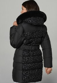 Donna che indossa un cappotto imbottito nero con pannelli leopardati e un ampio cappuccio rifinito in pelliccia, di profilo su uno sfondo grigio uniforme.