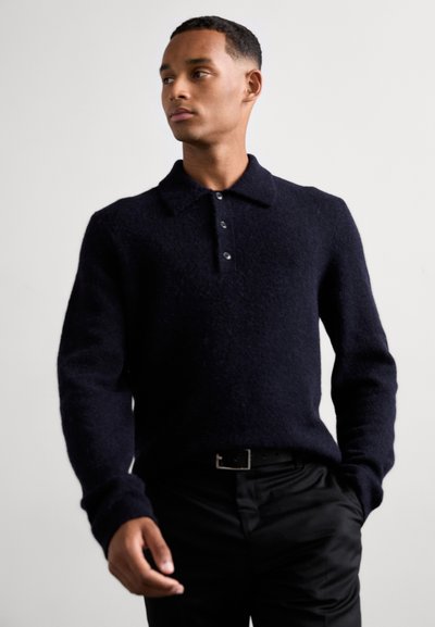 Marineblauer Strickpullover mit Polo-Kragen, der eine strukturierte Oberfläche und drei Knöpfe aufweist. Kombiniert mit schwarzen Hosen und einem Gürtel.