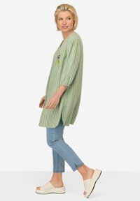 Camicia lunga a righe verdi con taschino frontale, abbinata a jeans azzurri chiari e sandali bianchi con zeppa. Vestibilità comoda e rilassata con maniche a gomito.