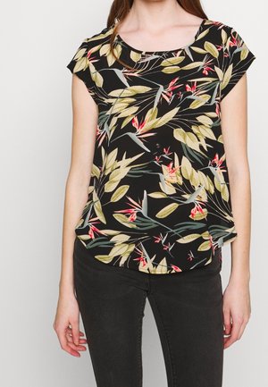 Femme portant un chemisier noir à manches courtes avec un motif de feuilles tropicales vertes et rouges, associé à un pantalon noir ajusté sur un fond uni.