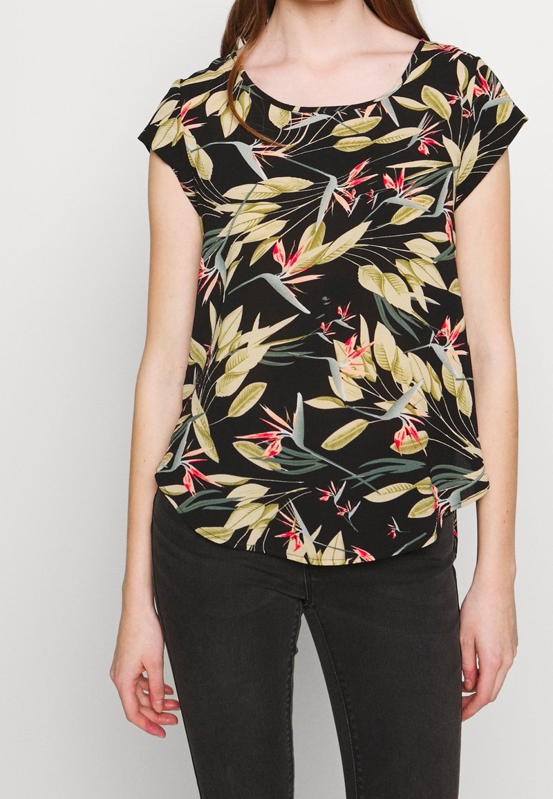 Femme portant un chemisier noir à manches courtes avec un motif de feuilles tropicales vertes et rouges, associé à un pantalon noir ajusté sur un fond uni.