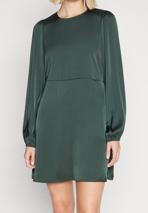 Femme portant une mini-robe à manches longues en satin vert foncé avec des poignets froncés et un col rond, debout devant un fond uni.