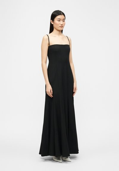 Femme debout, portant une longue robe noire sans manches avec de fines bretelles et des chaussures blanches pointues, sur un fond clair uni.