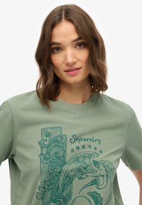 Camiseta de algodón en verde oliva con un diseño gráfico azul impreso que presenta elementos decorativos y el nombre de la marca "Superdry."