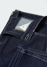 Jean en denim bleu foncé avec fermeture éclair partiellement ouverte, bouton fermé, et poche avant ainsi que la doublure de la poche intérieure visibles.