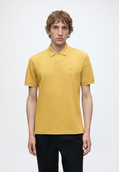 Jeune homme aux cheveux bouclés portant un polo jaune moutarde à manches courtes et un pantalon noir, debout devant un fond clair uni.