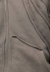 Grå zip-up jakke med strukturerede detaljer, der har en rå kantlomme og et blødt, bomuldsagtigt materiale. Enkelt, funktionelt design.