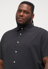 Chemise à manches courtes, à boutons, en tissu gris foncé. Présente un col rabattu et six boutons blancs sur le devant. Texture lisse.