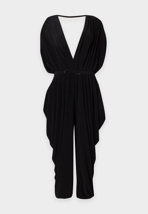 Høj kvinde med kort hår iført en sort jumpsuit med dyb V-hals og bælte, poserende med den ene hånd på hoften og sorte sandaletter med hæl.