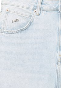 Lysblå denimjeanslomme med en lille metalnål med stiliseret tekst, synlige syninger og nitter.