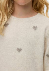 Bežna pletena jopica s krožnim vratom, ki vsebuje razpršene sivo srčne motive. Mehka tekstura in preprost dizajn izboljšata celoten videz.