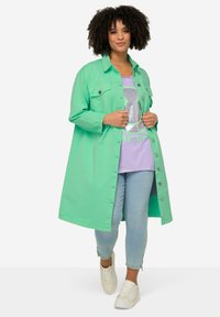 Giacca verde oversize con chiusura a bottoni, due tasche sul petto, indossata sopra una maglietta grafica viola e jeans skinny blu chiaro. Sneakers bianche.