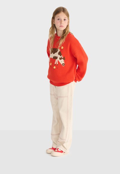 Jeune fille debout, portant un pull orange avec un motif de vache, un pantalon large crème et des chaussures rouges ornées de fleurs blanches.