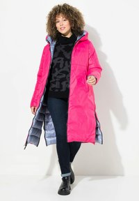 Abrigo acolchado rosa con un forro gris, que cuenta con capucha, cierre de cremallera, dos bolsillos laterales y un diseño hasta la rodilla. Se lleva sobre un suéter negro.