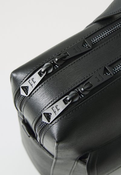 Black textured leather bag featuring dual metal zippers with triangular pulls. Minimalist design with subtle branding on zipper pulls.