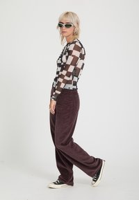 Personne avec des cheveux blonds courts portant un haut à manches longues à carreaux noir et blanc, un pantalon large marron foncé, des baskets noires et des lunettes de soleil blanches.