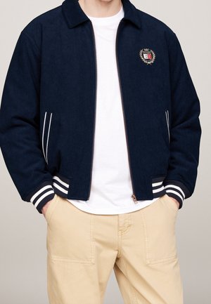 Mand iført en marineblå lynlåstrøje med hvide stribede ærmekanter og patch-logo, hvid T-shirt og beige bukser med hænderne i lommerne.