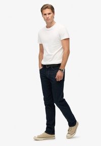 Camiseta blanca de algodón, jeans de mezclilla azul oscuro, zapatillas beige. Presenta un cinturón y un reloj. Diseño ajustado.