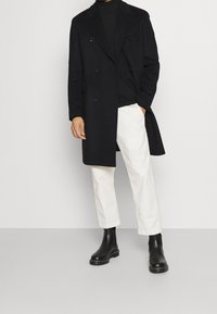 Sort dobbeltradet uldfrakke parret med en sort rullekrave, hvide bukser og sorte Chelsea-støvler. Glat tekstur og skræddersyet pasform.