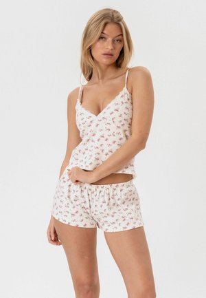 Nő, aki fehér pizsamát visel, kis rózsaszín virágos mintával, ujjatlan camisole felsővel és gumis derekú rövidnadrággal, egyszerű háttér előtt áll.