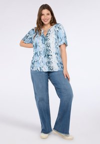 Top bluetto con motivo, maniche corte e scollatura a chiave, abbinato a jeans azzurri a gamba larga e scarpe bianche.