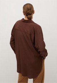 Chemise marron surdimensionnée avec un col, des manches longues et une texture de tissu lisse ; présentée de dos sur un fond clair.