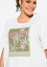 Camiseta blanca de gran tamaño con un gráfico botánico en verde y rosa que incluye la palabra "BLOOM" en letras negritas y diseños florales.