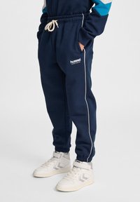 Marinblå atletiska joggers med elastisk midja, vit dragsko och sidofickor, med en logotyp och vit sidopiping. Matchas med vita sneakers.