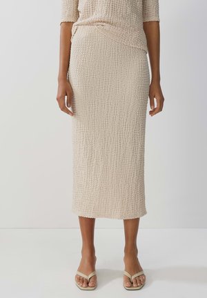 Knit beige midi skirt with textured, crinkled fabric, fitted silhouette, and a straight hem, worn with light flip-flops.