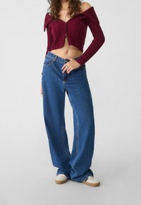 Bordeauxrode cropped trui met off-shoulder kabelgebreide structuur en geribbelde textuur, gecombineerd met losse blauwe spijkerbroek en witte sneakers.