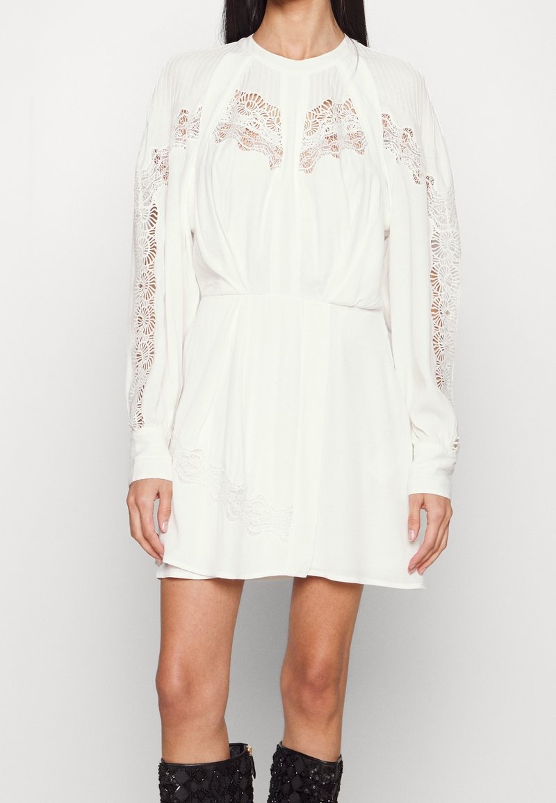 Femme portant une mini-robe blanche à manches longues avec des découpes en dentelle sur les manches, la poitrine et l'ourlet, associée à des bottes noires texturées montant jusqu'aux genoux.