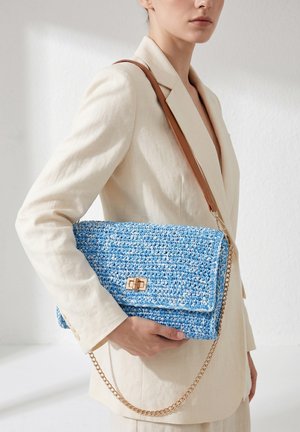 Femme en blazer crème tenant une pochette bleue tissée avec une lanière marron et un détail chaîne dorée.